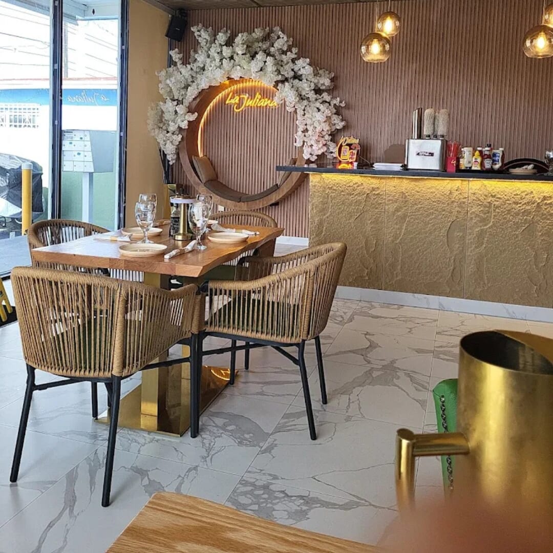 Commercial red-oak restaurant table on gold square base with stackable tan rope armchairs and floral La Juliana backdrop.
