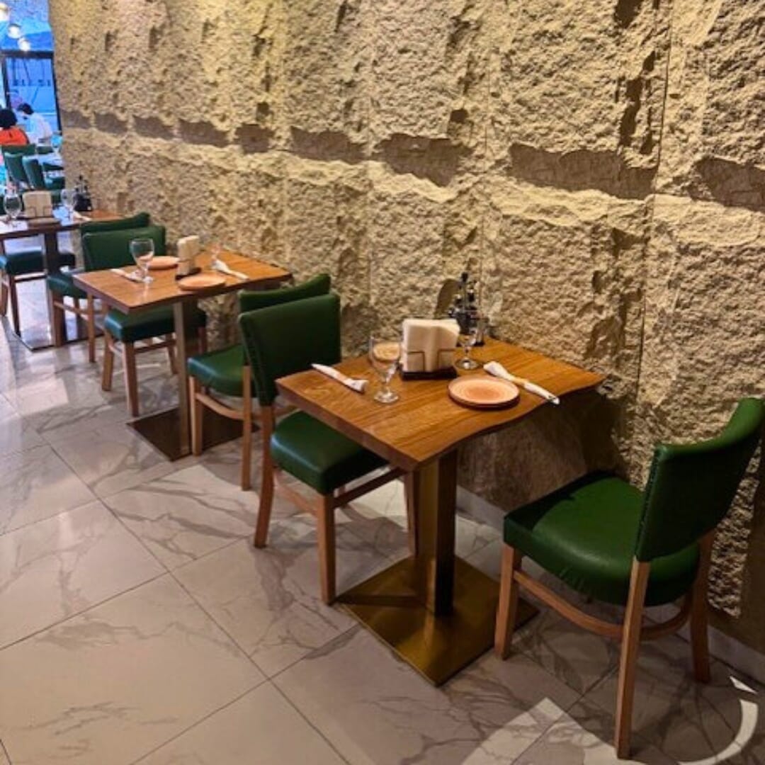Intimate two-top red-oak table on gold square base with green upholstered chairs against textured wall in La Juliana Miami.